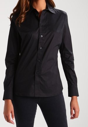 Woman wearing a fitted long-sleeve black button-up shirt and black pants, standing with arms relaxed by her sides.