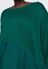 Zielony, oversize top z okrągłym dekoltem, asymetrycznym krojem oraz miękkiego materiału z delikatnym połyskiem. Bezszwowe miejsca zapewniają luźny fit.