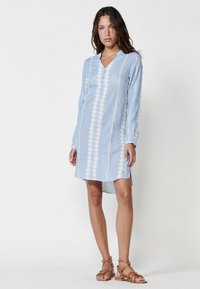 Helles blaues Tunika-Kleid aus strukturiertem Stoff, mit V-Ausschnitt, langen Ärmeln und weißen Stickereidetails entlang der Vorder- und Seitenansicht.