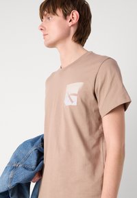 T-shirt en coton marron avec un col rond, des manches courtes et une petite impression graphique sur la poche ; le modèle tient une veste en denim.