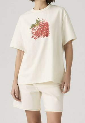 Kvinde iført en cremefarvet oversized t-shirt med en stor rød jordbærgrafik og matchende cremefarvede shorts mod en ensfarvet baggrund.