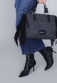 Borsa grigia con dettagli in pelle nera e frange, abbinata a stivali neri con tacco a punta. Pantaloni in denim visibili sotto.