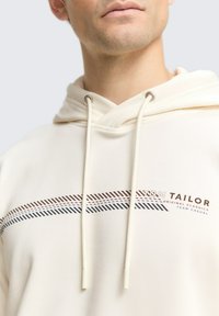 Beige Kapuzenpullover mit Kordelzug und grafischem Design in dunkelbraun, blau und roten Streifen über der Brust. Sanfte Textur.