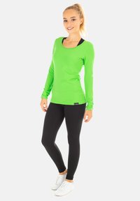 Grünes Langarmshirt kombiniert mit schwarzen Leggings und weißen Sneakern. Das Shirt hat einen runden Ausschnitt und ein schlankes Design.