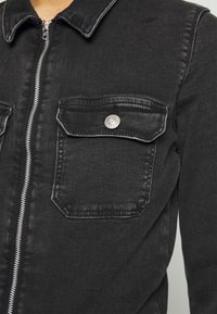 Veste en denim noir avec une fermeture éclair sur le devant, deux poches poitrine avec boutons-pression, et un col classique. Le tissu présente une légère texture.