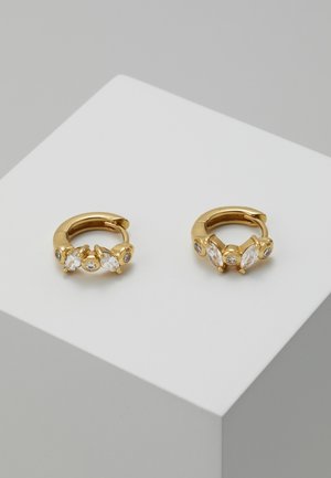 Gouden oorringen in de vorm van hoepels met elk drie heldere edelstenen, tentoongesteld op een wit geometrisch oppervlak tegen een grijze achtergrond.