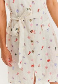 Lniana sukienka na kremowym tle, z intensywnymi, wielokolorowymi wzorami, paskiem w talii i drewnianymi guzikami, prezentująca swobodny styl.