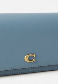Blå skinnlommebok med et gullfarget "C"-logo, glatt tekstur og kontrasterende sømmer langs kantene.