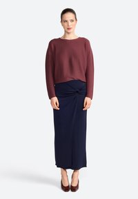 Pull en maille bordeaux avec un col rond, associé à une jupe bleu marine jusqu'au genou avec un détail torsadé à la taille. Talons bruns.