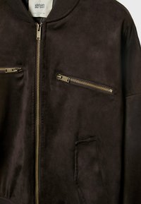 Veste en daim marron foncé avec fermeture éclair sur le devant et deux poches poitrine horizontales zippées, avec un petit col et des poignets côtelés.
