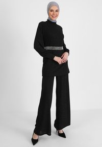 Outfit nero con un top lungo e strutturato e pantaloni a gamba larga, accentuato da una cintura con motivo. La modella indossa un foulard grigio e tacchi.