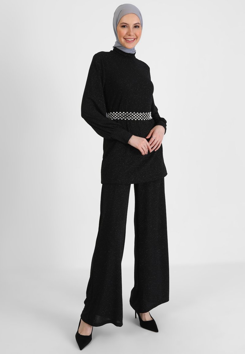 Outfit nero con un top lungo e strutturato e pantaloni a gamba larga, accentuato da una cintura con motivo. La modella indossa un foulard grigio e tacchi.