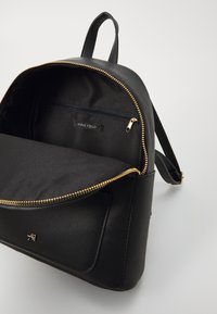 Sac à dos noir en matériau synthétique texturé avec une fermeture éclair dorée, comportant un grand compartiment principal et une petite poche intérieure.