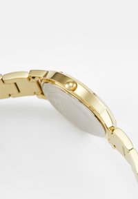 Montre en métal doré avec un bracelet à maillons, présentant un boîtier rond, une couronne texturée et un dos lisse et réfléchissant. Design simple et élégant.