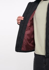 Chaqueta negra acolchada con forro burdeos, que presenta una cremallera y un diseño acanalado en el exterior, usada sobre una camisa verde claro.