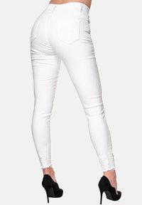 Jeans blancos ajustados con cintura alta, bolsillos traseros y detalle de dobladillo desgastado, combinados con zapatos de tacón negros.