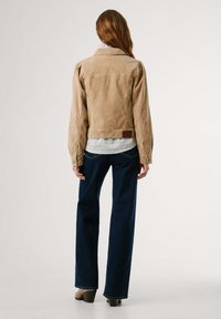 Khaki corduroy jas met een fitted ontwerp, schouderplooien en een bruine leren label op de taille, gedragen over een gestructureerd wit shirt en donkere broeken.