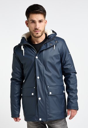Mann trägt eine marineblaue wasserdichte Jacke mit weißen Knöpfen und einer Kapuze mit weißem Fleece-Futter, steht vor einem einfarbigen Hintergrund.