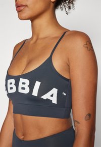 Pelēks sporta krūšturis ar plānām siksniņām, kurā ir lieli balti burti, kas veido vārdu "EBBIA", un atbilstoša lente apakšā. Gluda tekstūra.