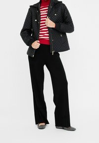 Chaqueta negra acolchada con capucha desmontable, combinada con un suéter de rayas rojas y blancas y pantalones negros de pierna ancha. Zapatos planos negros con patrón completan el conjunto.