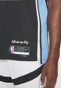 Musta koripallopaita, jossa on vaaleansinisiä yksityiskohtia, verkkokudostekstuuria ja NBA-logo. Sisältää tekstin "lift as we fly" ja kokolapun.