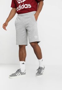 Man wearing gray Adidas shorts, white Adidas socks, gray Adidas sneakers, and a red Adidas T-shirt standing on a white background.