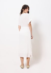 Weißes Midikleid mit kurzen Ärmeln, strukturiertem Stoff und gebundenem Taillenzug, verfügt über einen Seitenschlitz und wird mit beigen Schuhen kombiniert.