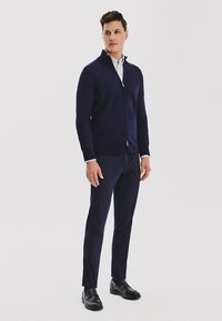 Granatowy sweter na zamek z gładkiego dzianiny, zestawiony z dopasowanymi ciemnymi spodniami i czarnymi mokasynami. Pod swetrem widoczna biała koszula z kołnierzykiem.