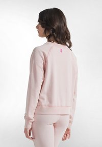 Sudadera de color rosa claro con puños y dobladillo acanalados. Presenta un cuello redondo y una pequeña etiqueta de logo rosa cerca de la parte trasera. Textura suave.