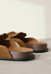Sandalias de ante marrón con puntera abierta, lados curvados y una suela de plataforma de madera gruesa y a rayas. Detalles texturizados y cosidos a lo largo de los bordes.
