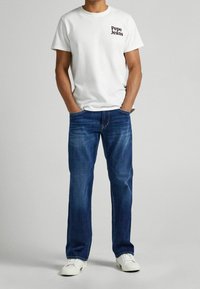 Camiseta de algodón blanca con el logo "Pepe Jeans" en el pecho, combinada con jeans de mezclilla azul. El calzado son zapatillas blancas. Diseño sencillo y casual.