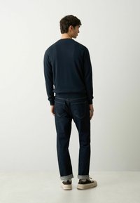 Marineblaue Sweatshirt mit Rundhalsausschnitt, kombiniert mit dunklen Jeans. Die Jeans haben einen umgeschlagenen Saum, und das Outfit wird mit hellen Turnschuhen abgerundet.