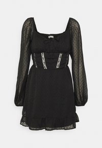 Black dress with long sheer polka dot sleeves, fitted bodice, lace accents, and a ruffled hem. Features a square neckline and tie detail.