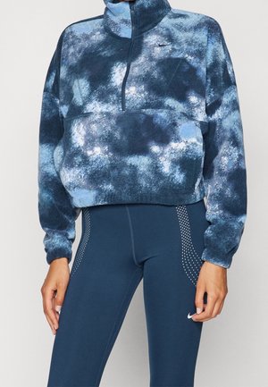 Vrouw draagt een blauwe en witte tie-dye fleece trui en marineblauwe leggings met witte gestippelde zijkanten en een klein logo op de dij.