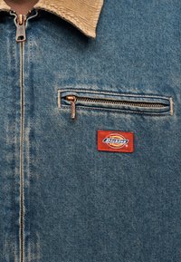 Μπουφάν τζιν με γιακά από ταμπά κορντόρι, τσέπη στο στήθος με φερμουάρ και κόκκινη και μπλε ετικέτα με το λογότυπο «Dickies». Χάλκινα διακοσμητικά στοιχεία.