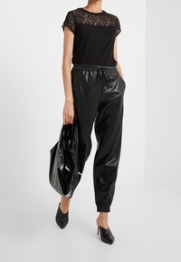 Mujer con top de encaje negro con mangas de capullo, joggers de cuero sintético negro, tacones de punta negra, llevando un gran bolso tote negro brillante.