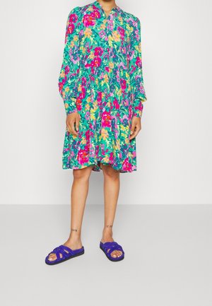 Blumenkleid in lebhaften Farben mit Grün, Pink und Gelb, langen Ärmeln und einem geknöpften Kragen. Kombiniert mit blauen Sandalen.