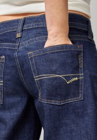 Temno modri trije denim hlače z rumenim šivom, ki vključujejo dva zadnja žepa in vidno roko v enem žepu. Posnetek z bližnjim kotom poudarja teksturo.