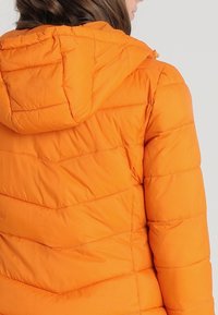 Orange puffjacka med huva, med quiltad chevron-sömnad och en slät, glansig yta. Bakre vy som visar fållen och sömmarna.