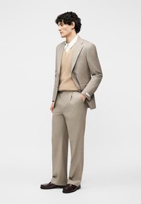 Άνδρας με μπεζ κοστούμι με λεπτές ρίγες, μπεζ γιλέκο, λευκό πουκάμισο και σκουριά καφέ παπούτσια, στέκεται με το ένα χέρι στην τσέπη μπροστά σε απλό φόντο.