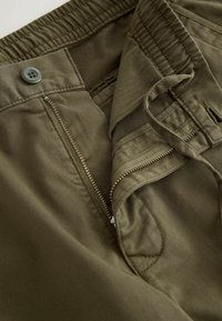 Olivgrüne Hose mit Reißverschluss und Knopfverschluss, ausgestattet mit einem elastischen Bund und Innenfächern. Weicher Baumwollstoff.