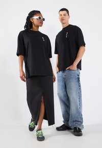 Deux mannequins portent des t-shirts noirs surdimensionnés. L'un porte une jupe en denim noir avec une fente avant. L'autre porte un jean clair ample et des baskets noires.