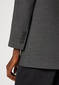 Manga de blazer gris con cinco botones negros, textura suave, diseño estructurado y puño ligeramente estrechado.