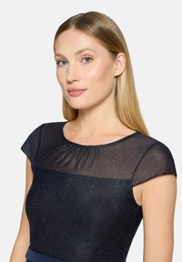 Robe bleu marine avec de courtes manches chauve-souris translucides et un col froncé. Le tissu présente un léger scintillement, créant un effet superposé élégant.