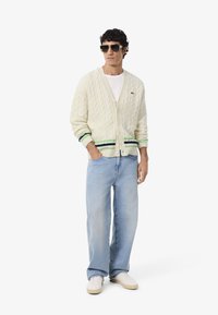 Cardigan a treccia beige con strisce verdi e blu navy all'orlo, abbinato a jeans azzurri e sneaker bianche. Occhiali da sole quadrati indossati.