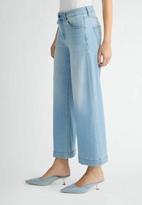 Jeans en denim bleu clair à jambes larges, avec une taille haute, des ourlets roulés et un léger effet délavé. Associés à des mules bleues ornées.