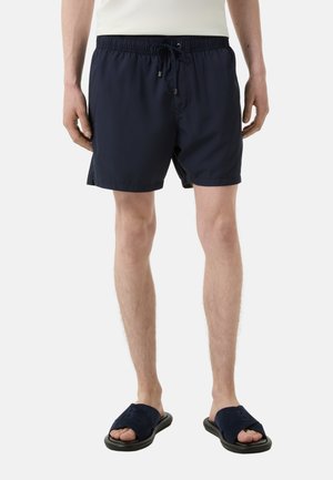 Marinefarbene Badeshorts mit elastischem Bund, Kordelzug und Seitentaschen; kombiniert mit marinefarbenen Slippern, die ein Logo am Riemen tragen.