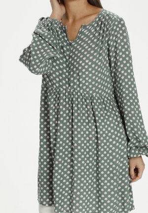 Kobieta ubrana w zieloną sukienkę z długim rękawem, z małymi białymi kwiatowymi wzorami i marszczoną talią, pozująca z jedną ręką przy twarzy.