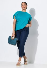 Tyrkysový krátký rukáv s volánkovými rukávy, tmavé džínové kalhoty s cvočkovými manžetami a tyrkysová kabelka s třásněmi a dřevěnou rukojetí.