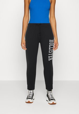 Person wearing black drawstring sweatpants with white "Hollister" text on leg, blue sleeveless top, and black high-top sneakers with white laces.